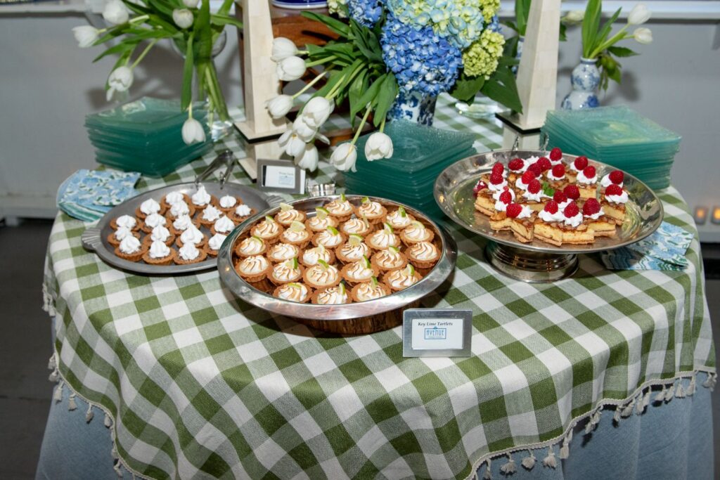 Three trays of assorted desserts rest on a green and white checkered tablecloth, surrounded by blue and white floral arrangements and stacked green napkins—a perfect setting for those seeking inspiration for engagement party venues in Atlanta.