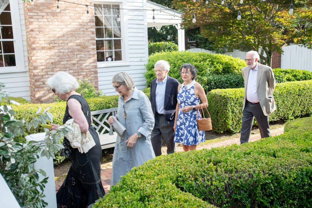 Five older adults walk in a line along a garden path outdoors, dressed in semi-formal attire, with greenery and a white building in the background—a scene reminiscent of gatherings at charming engagement party venues Atlanta has to offer.