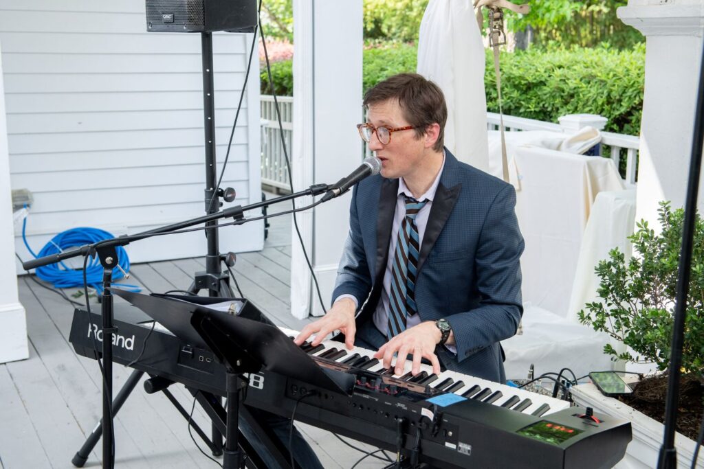 A man in a suit and tie plays a Roland keyboard and sings into a microphone on a porch. Surrounded by greenery and music equipment, the scene evokes the lively atmosphere often found at engagement party venues in Atlanta.