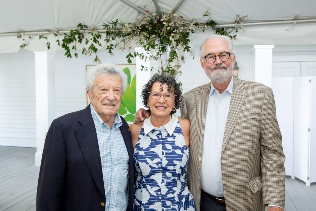Three older adults, two men and one woman, stand together smiling under a white tent with greenery above—an elegant setting reminiscent of top engagement party venues Atlanta has to offer. The men wear jackets and shirts, while the woman wears a blue and white sleeveless dress with patterns.