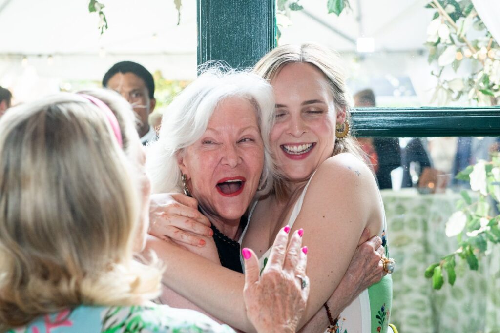 Two women, one older with white hair and one younger with blonde hair, are hugging and smiling joyfully at a lively outdoor event—perhaps hosted at one of the charming engagement party venues Atlanta has to offer—while another woman stands nearby.