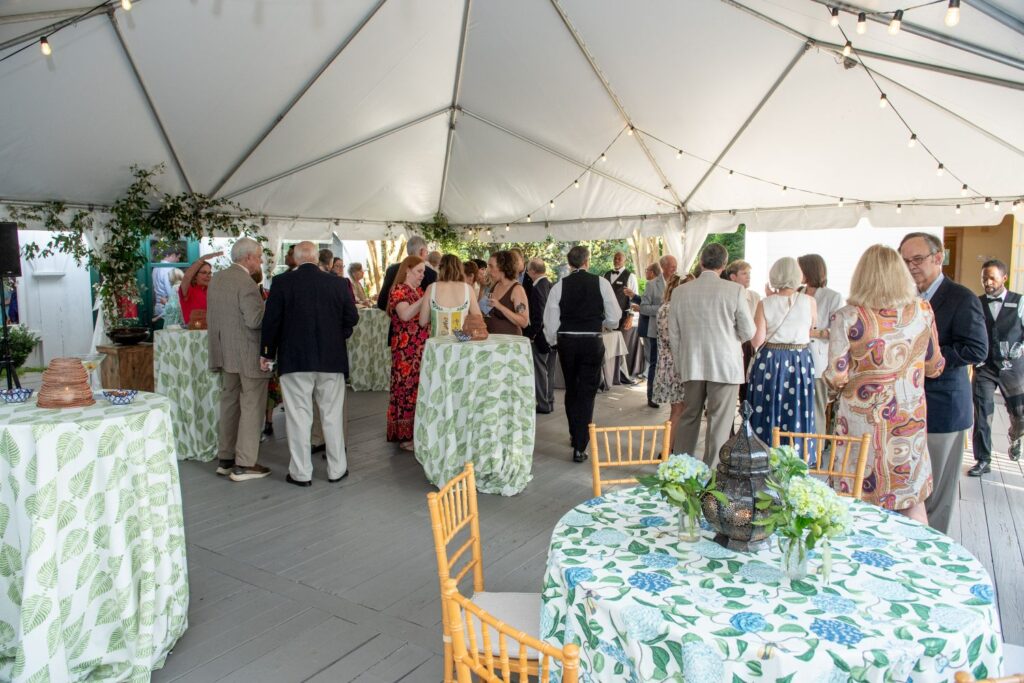 Guests mingle under a white tent at a formal event, reminiscent of the elegant gatherings found at top engagement party venues Atlanta offers. Round tables with floral tablecloths and green centerpieces are scattered around, while string lights hang overhead, creating a festive atmosphere.