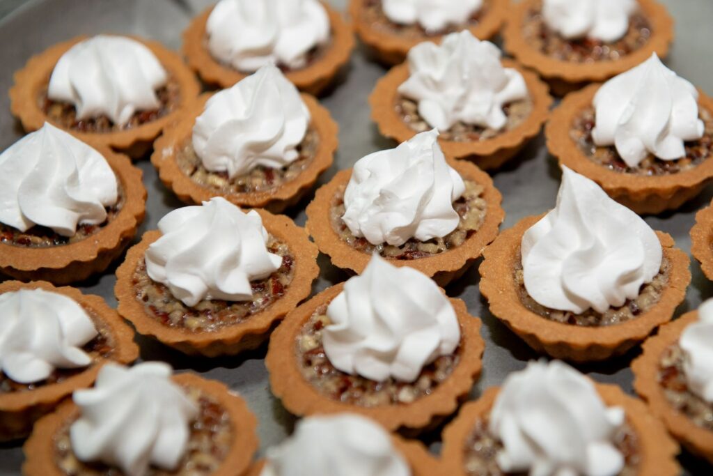 Mini pecan tarts arranged in rows, each topped with a generous swirl of whipped cream—perfect for serving at elegant engagement party venues Atlanta. The tarts have golden brown crusts and nutty filling visible beneath the cream.