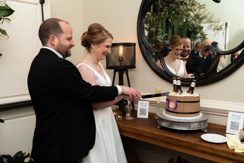A bride and groom smile as they cut a cake shaped like a wooden bucket with beer bottles, celebrating their special day at one of the charming wedding venues Atlanta has to offer. Their reflection appears in a round mirror behind them, while a QR code and stylish decor adorn the wooden table beside the cake.