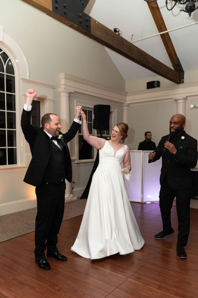 A bride and groom celebrate on the dance floor at one of the beautiful wedding venues Atlanta has to offer, smiling with raised arms. A man in a suit stands nearby, holding a microphone. The setting is an elegant indoor venue.