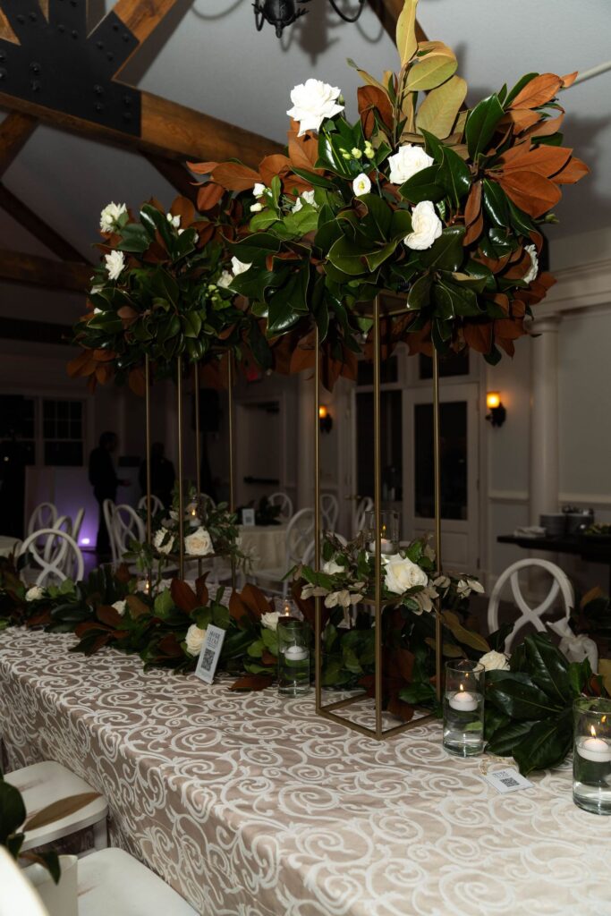 A long table with a patterned beige tablecloth features tall floral centerpieces with white roses and green leaves, surrounded by candles in glass holders in an elegant indoor setting—capturing the sophisticated ambiance found at some of the finest wedding venues Atlanta has to offer.