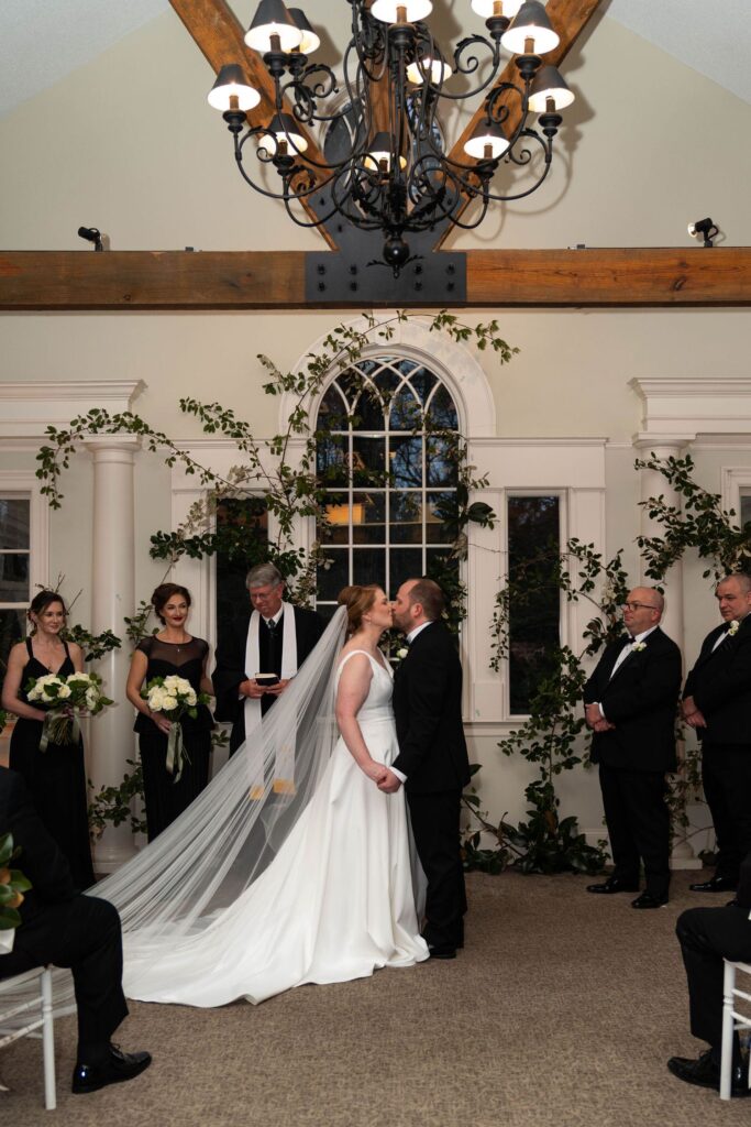 A bride and groom kiss at their indoor wedding ceremony, surrounded by the wedding party and officiant, under a chandelier, with greenery and white flowers as decor—a romantic moment that beautifully showcases why wedding venues Atlanta are renowned for their elegance.