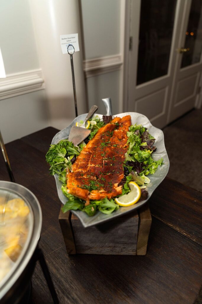 A platter of seasoned baked salmon on a bed of mixed greens, garnished with lemon wedges and herbs, with serving utensils nearby—a perfect choice for catering menus at wedding venues Atlanta couples adore.