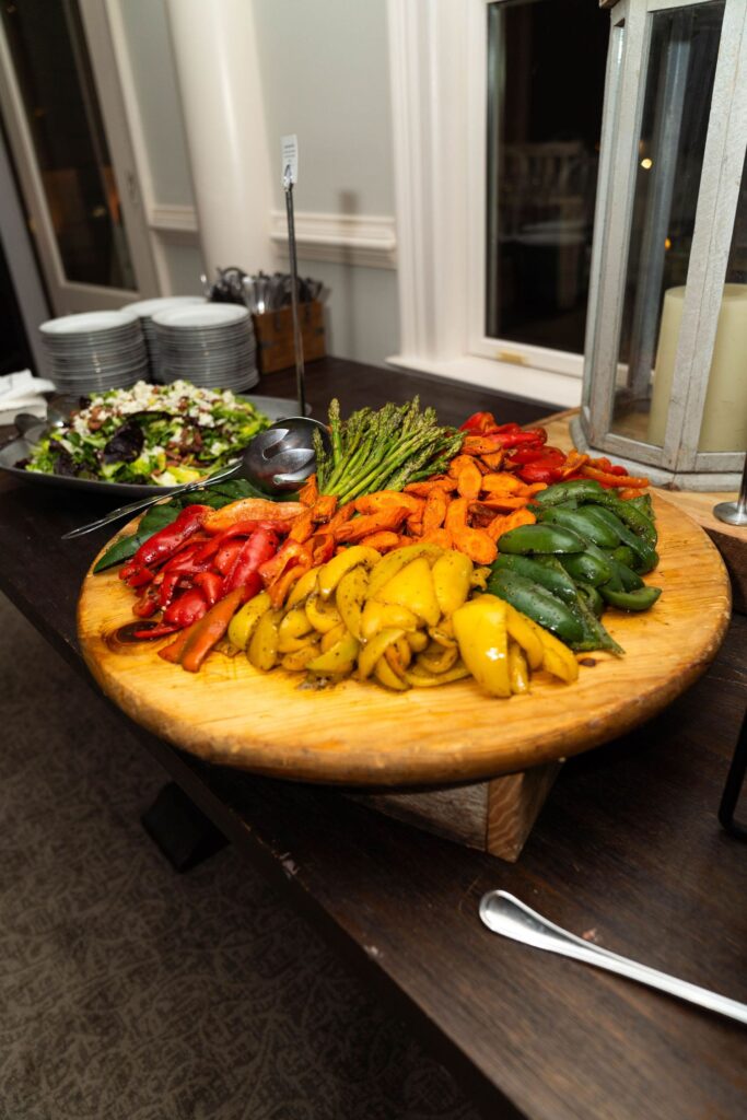 A wooden platter with colorful grilled vegetables—red, yellow, and green peppers, asparagus, and carrots—on a table next to a salad bowl, plates, and cutlery sets the scene for an elegant gathering at one of the charming wedding venues Atlanta has to offer.