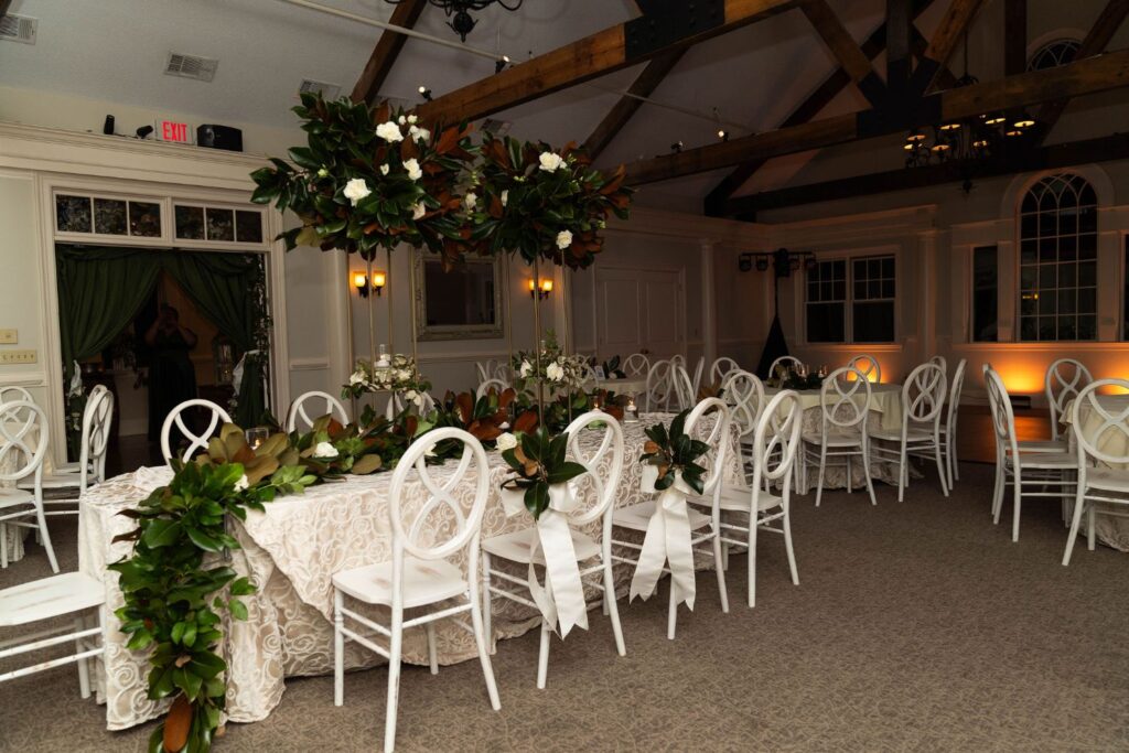Elegant banquet hall with white chairs, long tables covered in white cloth, and lush green foliage centerpieces. Wooden beams and chandeliers are visible on the ceiling. As one of the premier wedding venues Atlanta has to offer, the room is softly lit and empty of people, providing a serene setting for your special day.