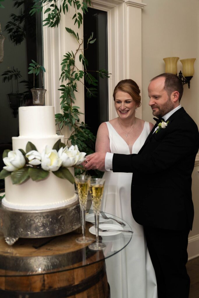 A bride and groom smile as they cut a white, three-tier wedding cake decorated with white flowers at one of the most elegant wedding venues Atlanta has to offer. Two champagne glasses sit on the table in front of them.