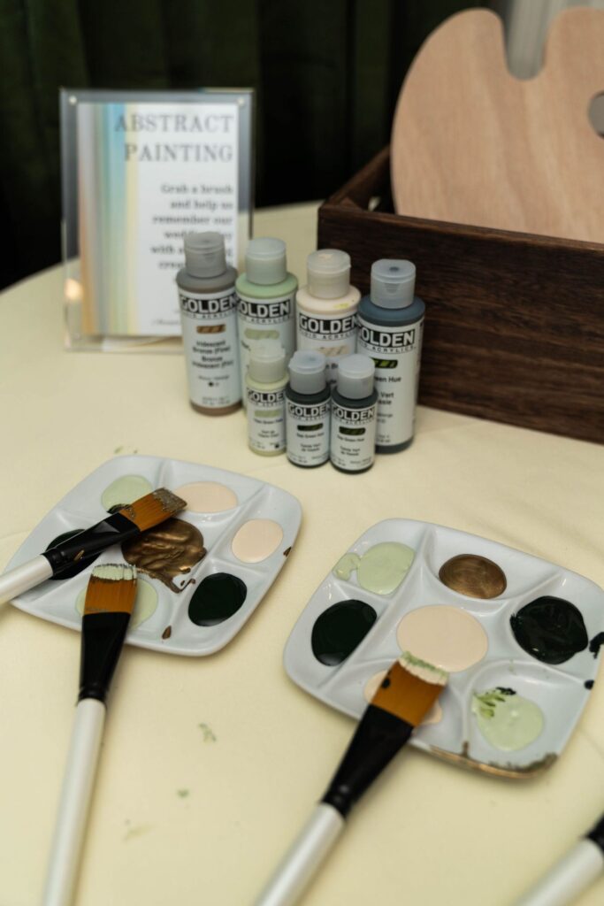 Art supplies on a table: paintbrushes, paint trays with assorted green, gold, and beige paints, bottles of Golden acrylic paint, a wooden palette, and a sign about abstract painting in the background—creating an atmosphere as inspiring as some of the most unique wedding venues Atlanta has to offer.