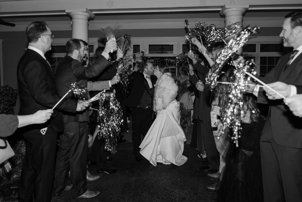 A bride and groom kiss while guests cheer and wave pom-poms during their nighttime wedding exit outside a building—one of the most memorable wedding venues Atlanta has to offer. The image is in black and white.