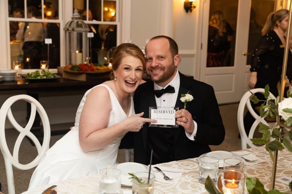 A bride and groom sit smiling at a decorated table, with the bride pointing at a Reserved sign held by the groom. The background—showcasing food on a buffet table and warm ambient lighting—reflects the elegant charm often found at top wedding venues Atlanta has to offer.