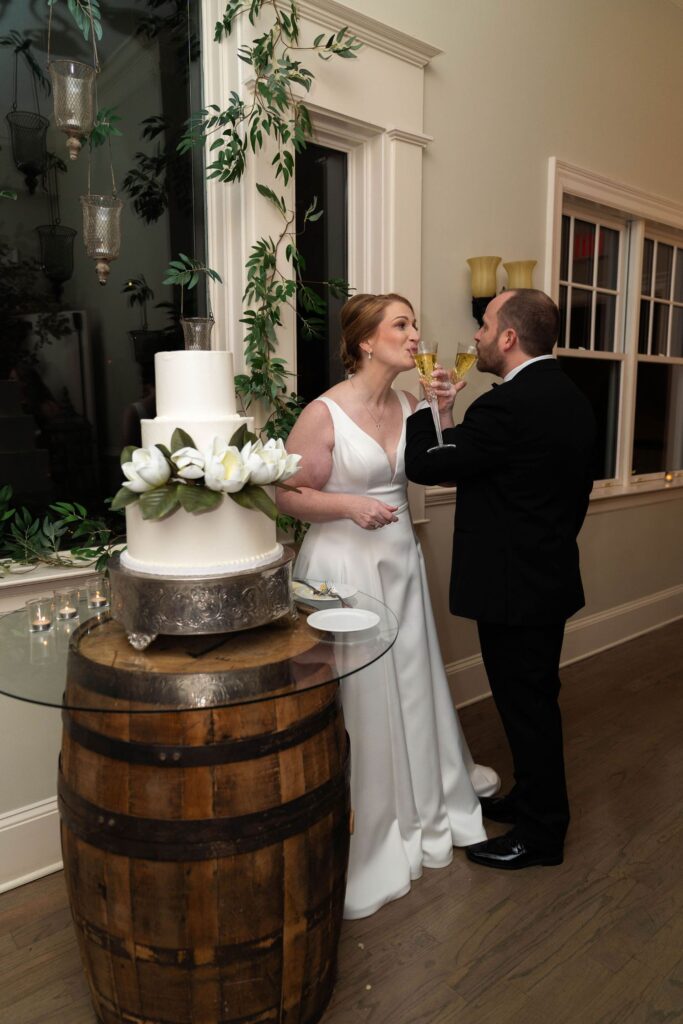 A bride and groom in formal attire share a toast beside a three-tiered white wedding cake on a wooden barrel at an indoor reception, showcasing the elegant atmosphere found in top wedding venues Atlanta has to offer.