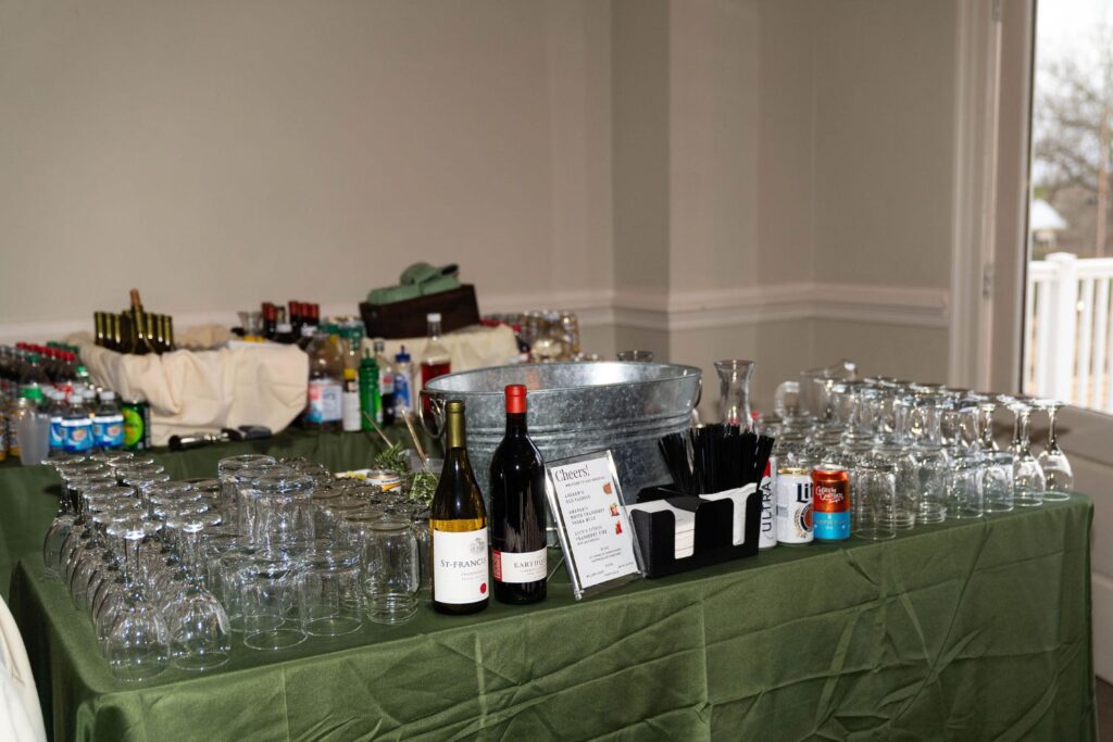 A table with wine, liquor bottles, cans, water, glassware, an ice bucket, and black straws on a green tablecloth, set up as a self-serve bar in a bright room near a window—perfect for elegant gatherings at top wedding venues Atlanta has to offer.