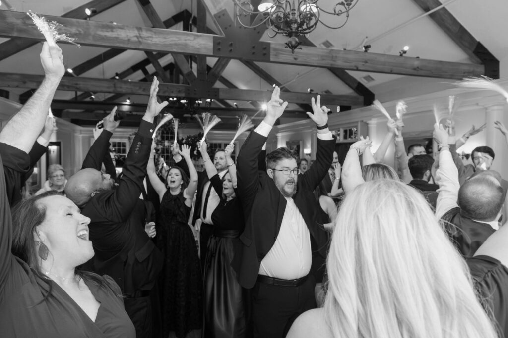 A group of people dressed formally celebrate indoors at one of the elegant wedding venues Atlanta has to offer, raising their arms and cheering with excitement under wooden beams and a chandelier.