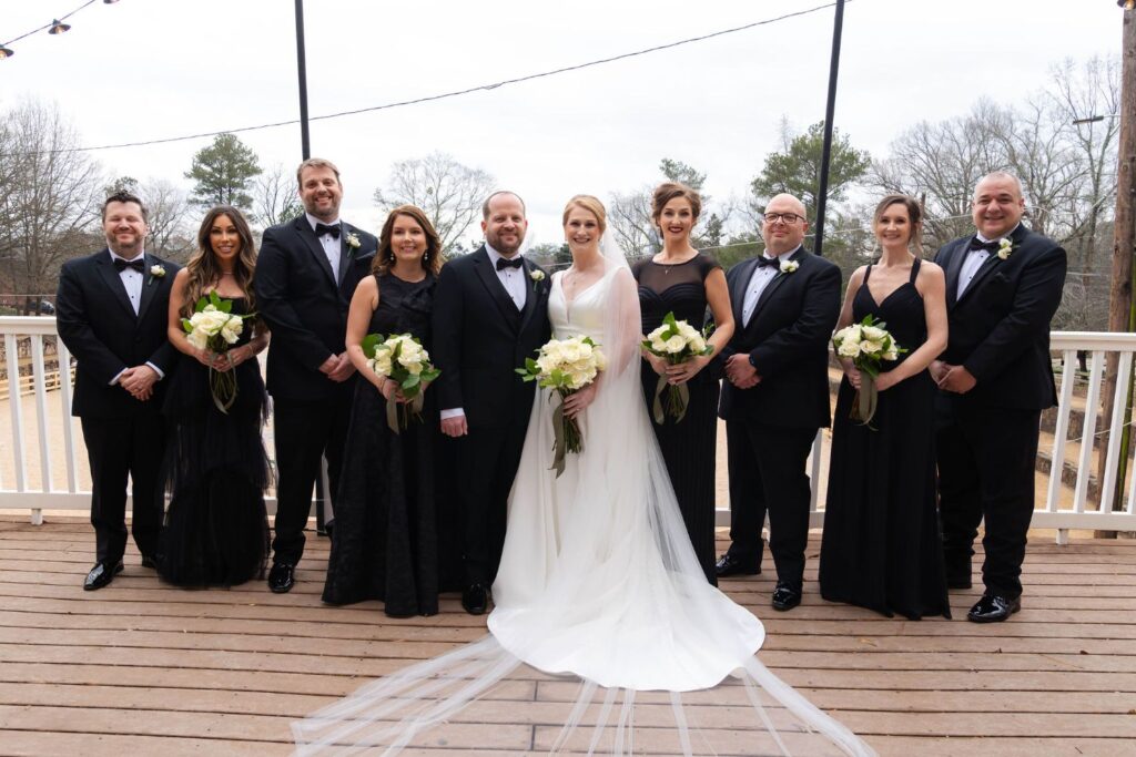 A wedding party poses on a wooden deck at one of the picturesque wedding venues Atlanta has to offer. The bride in white stands with the groom and eight attendants dressed in black, all holding bouquets and smiling at the camera. Trees and string lights are in the background, adding charm to this special celebration.