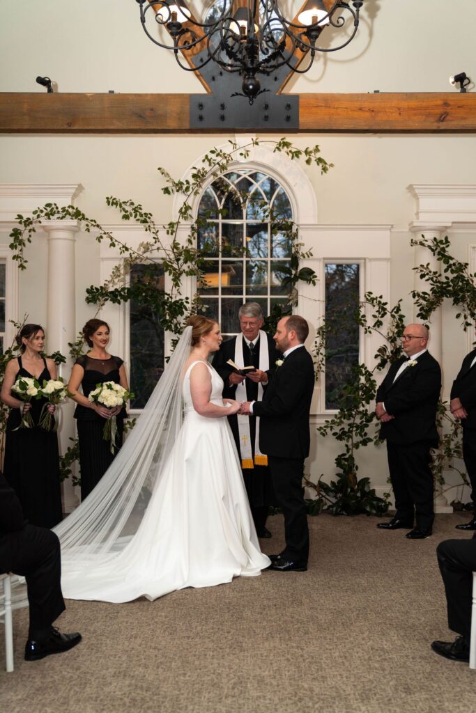 A bride and groom stand facing each other at an indoor wedding ceremony, holding hands as an officiant reads. Bridesmaids and groomsmen stand nearby, while lush greenery decorates the window behind them—showcasing the elegant charm often found at wedding venues in Atlanta.