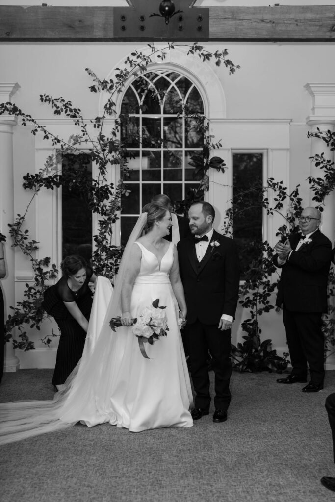 A bride and groom stand together indoors at one of the beautiful wedding venues Atlanta has to offer, holding hands and smiling after their wedding ceremony, with floral decorations and a large arched window behind them. A few guests look on, one taking a photo.