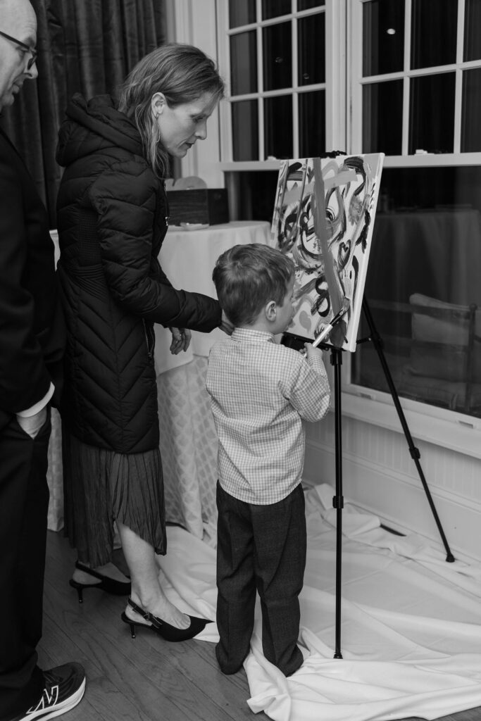 A woman helps a young boy paint on a canvas set on an easel indoors, with an adult standing nearby. The scene appears casual, reminiscent of candid moments found at elegant wedding venues in Atlanta, and the image is in black and white.
