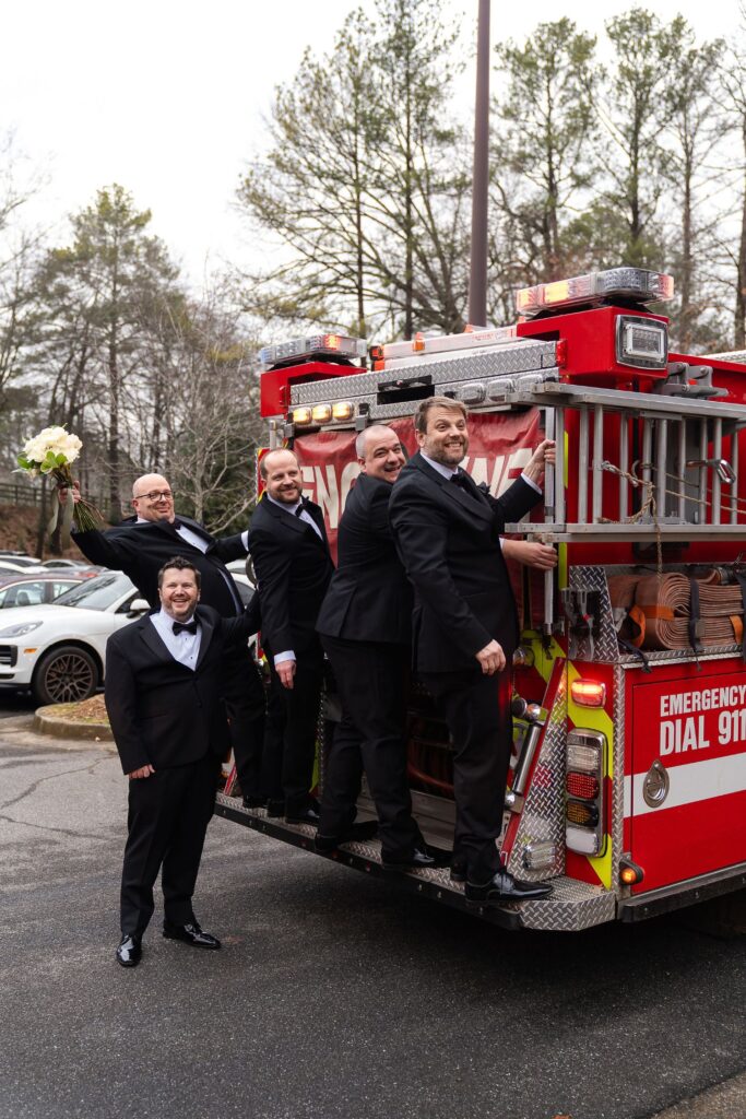 Five men in tuxedos, four standing on the back of a fire truck and one beside it, pose and smile for a playful photo outside on a cloudy day—capturing a fun moment at one of the unique wedding venues Atlanta has to offer.