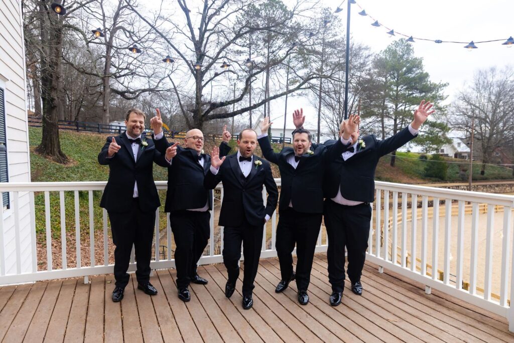 Five men in black tuxedos stand on a wooden deck at one of the most charming wedding venues Atlanta has to offer, four of them with arms raised and making excited expressions, while the man in the center stands calmly. Trees and string lights are visible in the background.