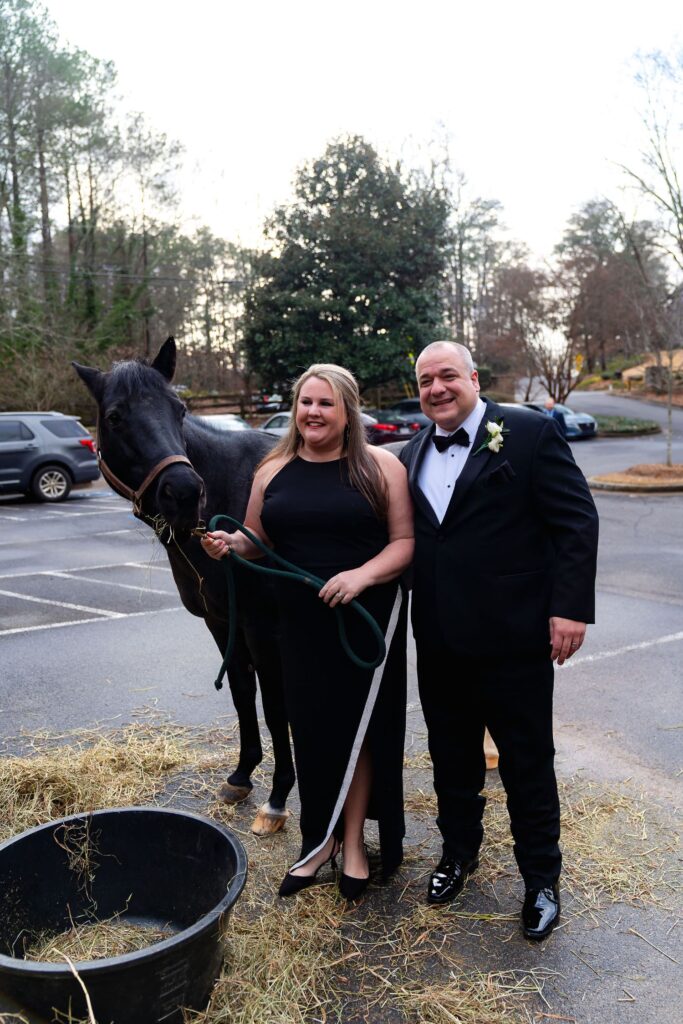 A woman in a black dress and a man in a tuxedo stand smiling next to a black horse in a parking lot with trees and parked cars in the background, capturing the elegance often seen at top wedding venues Atlanta has to offer.