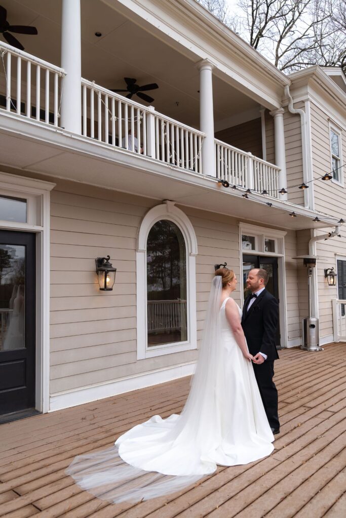 A bride in a white gown and veil holds hands with a groom in a black suit, standing on a wooden deck outside a large, light-colored building with a balcony and tall windows—one of the elegant wedding venues Atlanta has to offer.