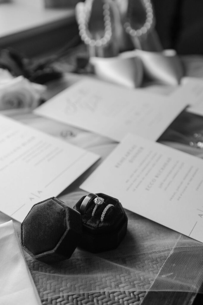 Black and white photo of wedding invitations, a rose, high-heeled shoes with pearls, and an open ring box displaying an engagement ring and two wedding bands on a woven surface—capturing the elegant details often found at top wedding venues Atlanta couples adore.
