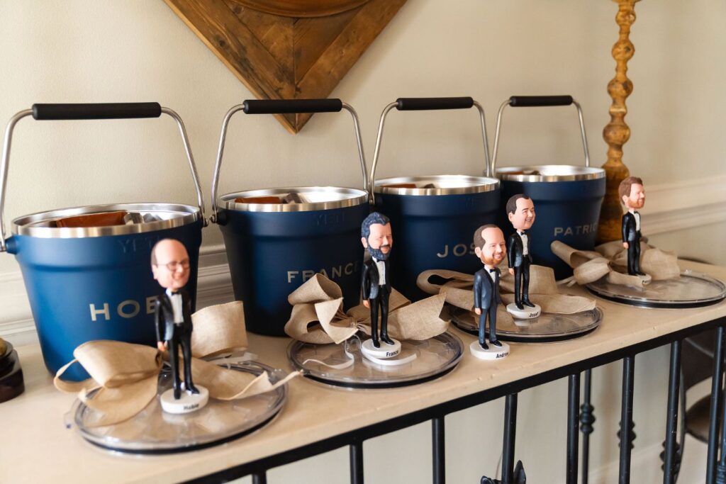 Five blue metal buckets with white engraved names sit on a shelf, each paired with a suited bobblehead figurine and a beige ribbon. The display appears organized and decorative—an inspiring touch often seen at top wedding venues Atlanta couples adore.