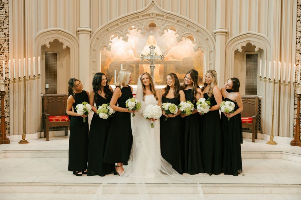 Seven women in black dresses and one bride in white stand smiling, holding bouquets, inside a church with a large painting and lit candles behind them—a beautiful moment before they head to one of the top wedding reception venues Atlanta has to offer.