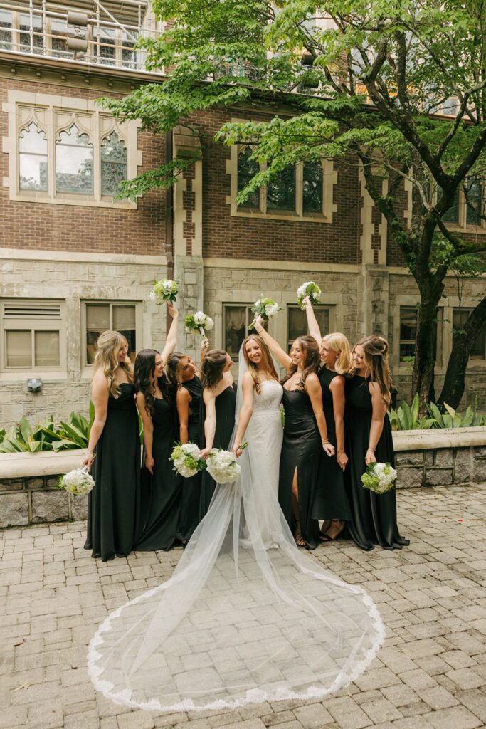 A bride in a white gown stands surrounded by bridesmaids in black dresses, holding bouquets and smiling, with trees and a brick building—one of the charming wedding reception venues Atlanta offers—in the background.