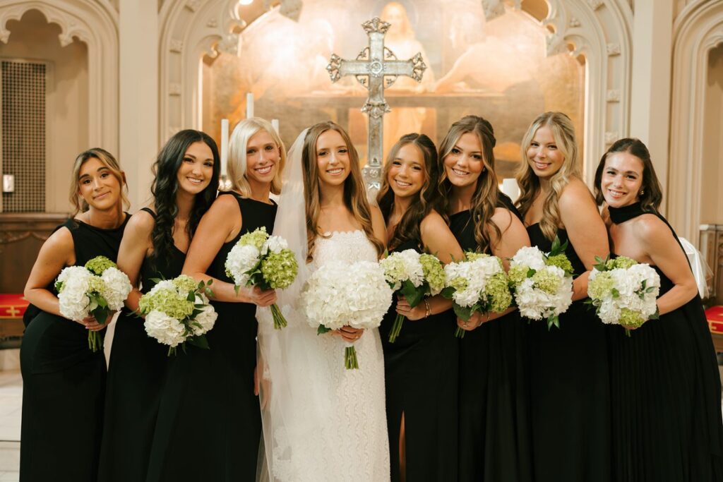 A bride in a white dress and veil stands with seven bridesmaids in black dresses, all holding white floral bouquets, smiling inside a church with a large cross in the background—a timeless moment before celebrating at one of the elegant wedding reception venues Atlanta has to offer.