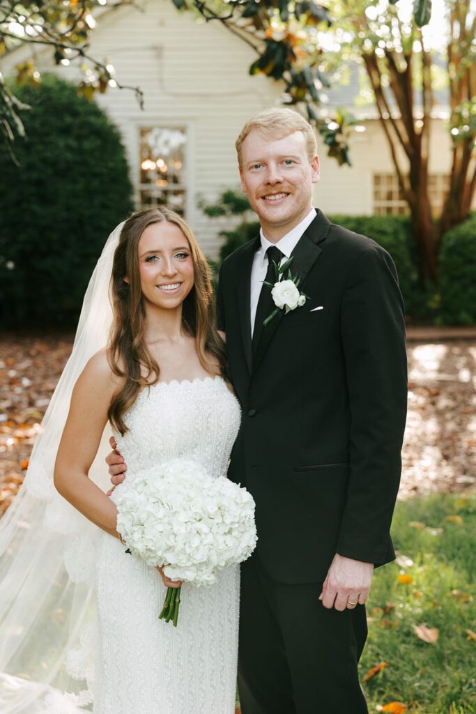 A bride in a white dress holding white flowers stands next to a groom in a black suit and tie, both smiling outdoors with greenery and a white building in the background, capturing the elegance found at some of the top wedding reception venues Atlanta has to offer.