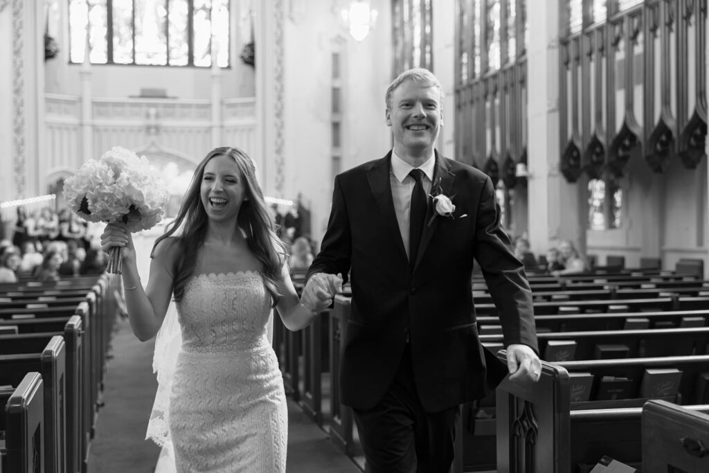 A bride and groom walk joyfully down the aisle of a church, holding hands and smiling, surrounded by pews and stained glass windows, their hearts filled with anticipation for the celebration ahead at one of the beautiful wedding reception venues Atlanta has to offer.