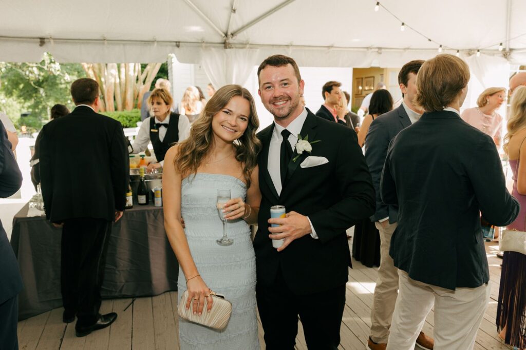 A woman in a light blue dress and a man in a black suit smile at a formal event under a tent, surrounded by other guests. She holds a drink and small clutch; he holds a can. The elegant atmosphere highlights why this location stands out among top wedding reception venues Atlanta has to offer.