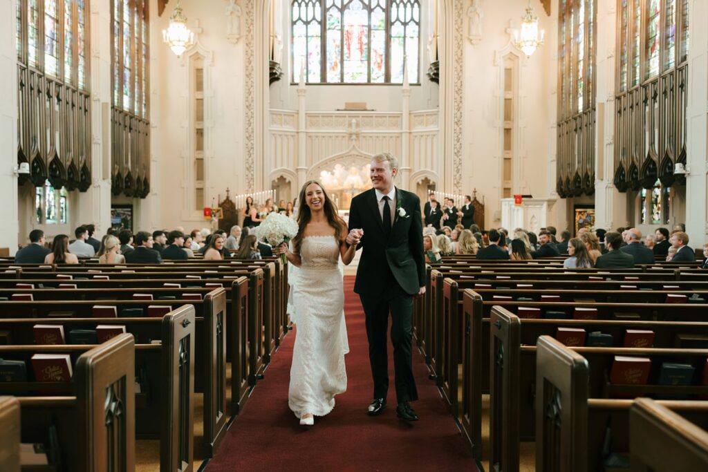 A bride and groom smile as they walk down the aisle together in a church filled with seated guests. The bride holds a bouquet and wears a white dress; the groom wears a black suit. Stained glass windows decorate the background, evoking the elegance found in top wedding reception venues Atlanta has to offer.