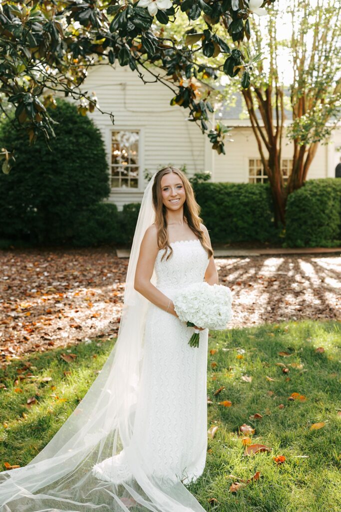 A bride in a white dress and long veil holds a bouquet of white flowers, standing on grass with trees, sunlight, and a house—one of the elegant wedding reception venues Atlanta has to offer—in the background.