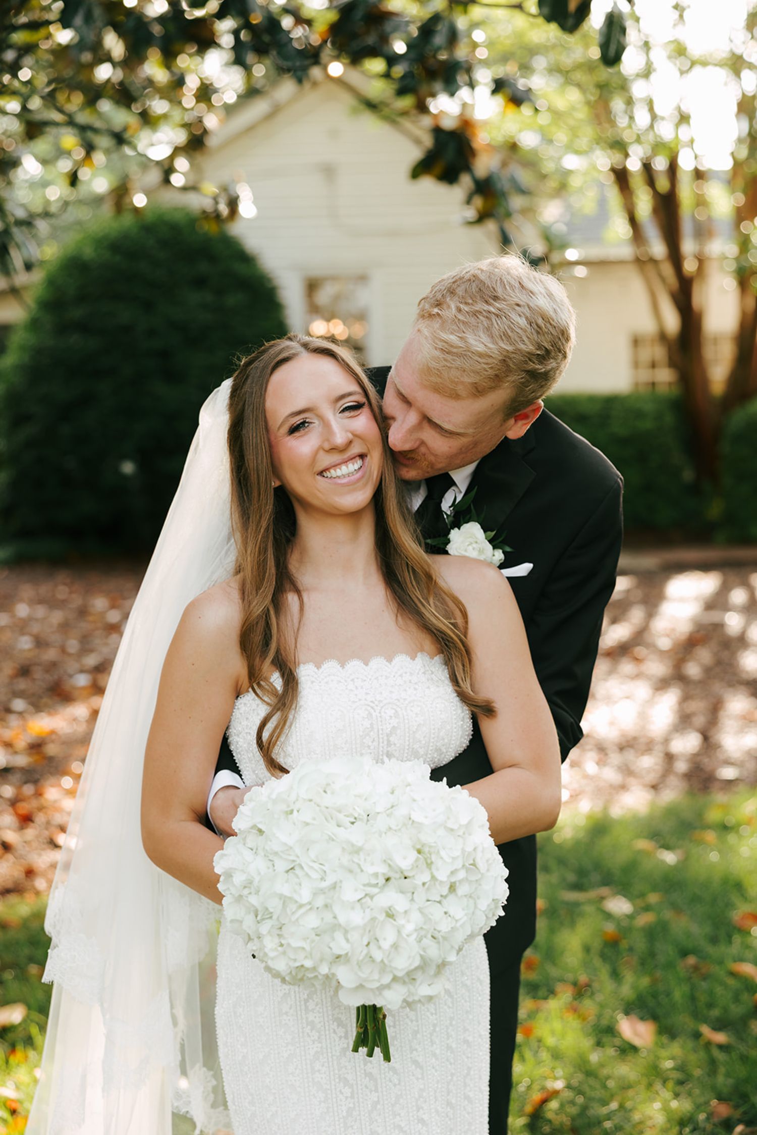 A bride in a white dress holding a bouquet of white flowers smiles while a groom in a black suit hugs her from behind and kisses her head in a sunlit outdoor garden setting, capturing the joy often found at top wedding reception venues Atlanta has to offer.