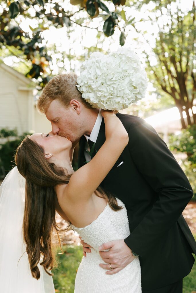 A bride and groom share a kiss outdoors at one of the stunning wedding reception venues Atlanta has to offer. The bride holds a bouquet of white flowers and wears a veil, while the groom wears a black suit. Sunlight filters through the trees in the background.