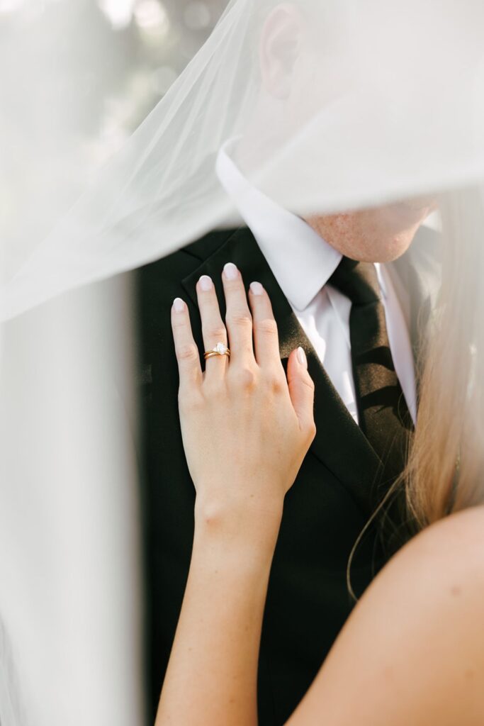 A bride gently rests her hand, showing an engagement ring, on a groom’s chest. Both are partially covered by a sheer white veil, with only their lower faces visible. The groom wears a black suit and tie—capturing an intimate moment often cherished at top wedding reception venues Atlanta has to offer.