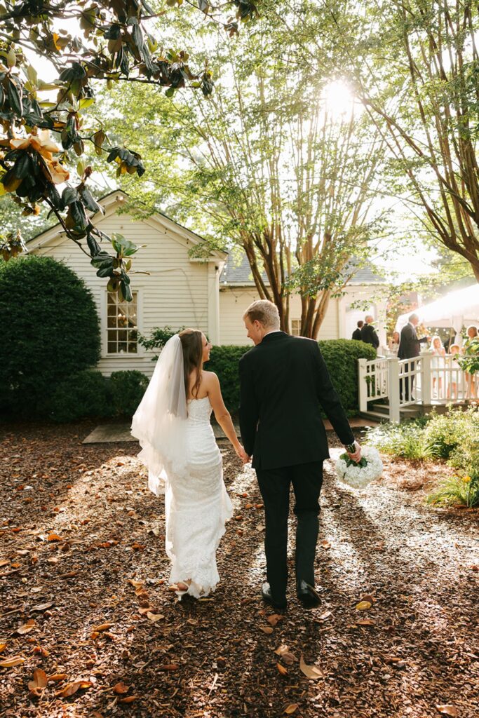 A bride and groom walk hand-in-hand outside, with sunlight filtering through trees at one of the charming wedding reception venues Atlanta has to offer. The bride wears a white dress and veil, while the groom is in a dark suit, holding a bouquet. Guests gather in the background near a white house.