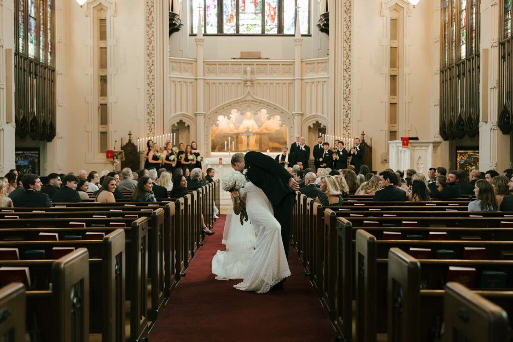 A bride and groom kiss in the aisle of a church filled with guests. Bridesmaids and groomsmen stand at the front, while sunlight streams through stained glass windows above the altar—a moment that captures the timeless beauty often sought after when exploring wedding reception venues Atlanta has to offer.