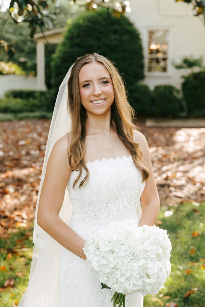 A woman in a white wedding dress and veil stands outside holding a bouquet of white flowers, smiling at the camera with greenery and one of the elegant wedding reception venues Atlanta has to offer in the background.