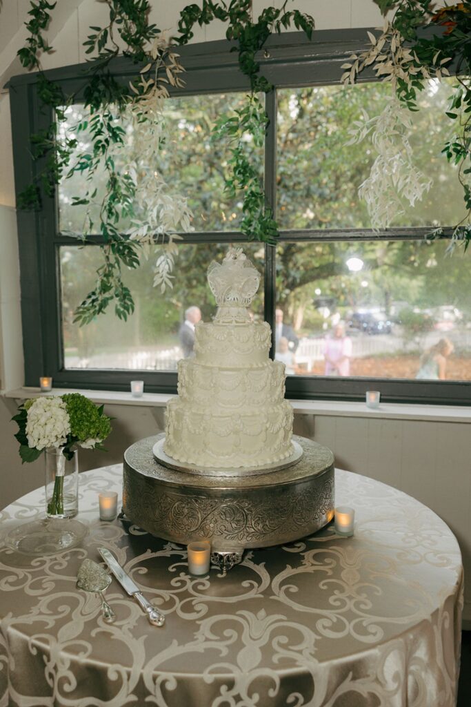 A white tiered wedding cake with floral decorations sits on a silver stand atop a round table with a patterned cloth, surrounded by candles and flowers, in front of a window with greenery hanging above—creating an elegant scene that captures the charm found at top wedding reception venues in Atlanta.