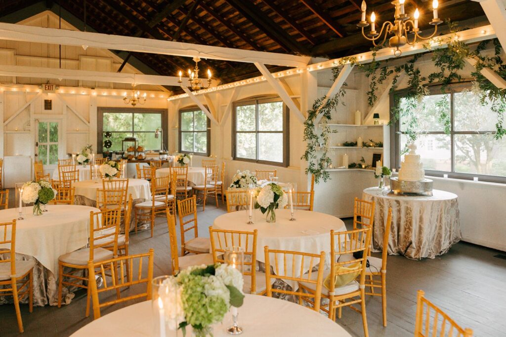 A bright, rustic wedding reception hall—one of the most charming wedding reception venues Atlanta has to offer—features wooden beams, round tables with white cloths, gold chairs, floral centerpieces, string lights, and a decorated cake table by large windows.