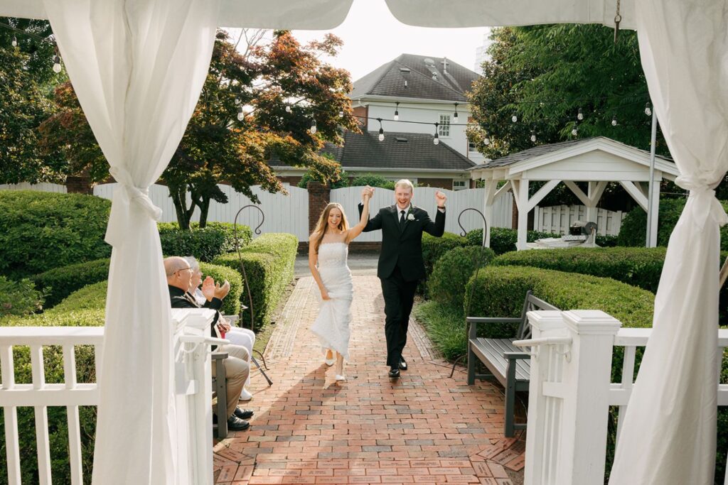 A bride and groom walk arm in arm down a brick path, smiling and raising their hands in celebration as guests seated on the left applaud. The scene is outdoors with green hedges and a white gazebo, showcasing the charm often found at top wedding reception venues Atlanta has to offer.