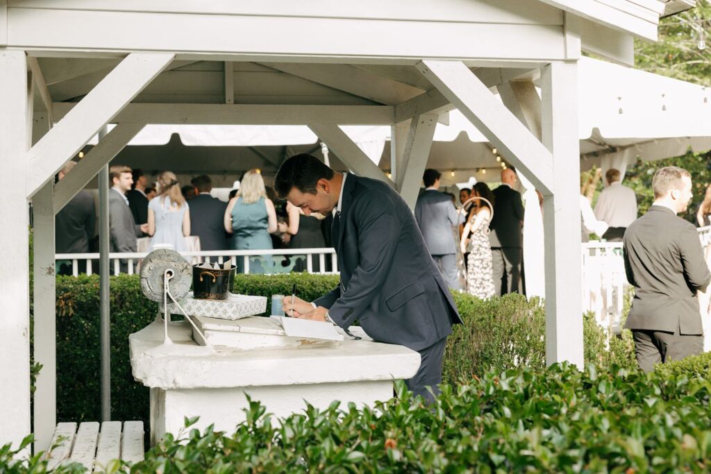 A man in a suit signs a book on a stone well under a gazebo, with guests gathered in the background at an outdoor event, capturing the elegant atmosphere often found at top wedding reception venues Atlanta has to offer.