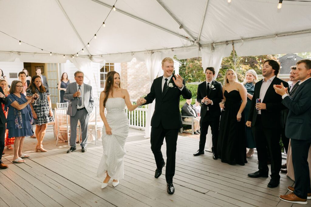 A bride and groom enter a tented outdoor reception, holding hands and smiling, as guests in formal attire watch and celebrate. String lights hang overhead, people are taking photos and clapping—all capturing the magic found at wedding reception venues Atlanta couples adore.